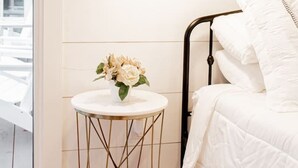 A bedroom with a white bed, a bedside table with a vase of flowers, and a wall-mounted lamp.