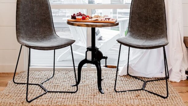 A room with two chairs, a table with a fruit platter, a hanging light fixture, and a view through a window with a curtain.