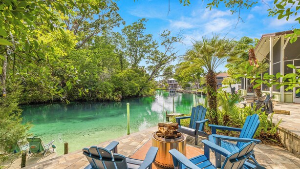 A patio with outdoor seating, a fire pit, and a view of a river surrounded by greenery. - Stay directly on crystal clear waters of Weeki Wachee River with your own beach! (weekiwachee)