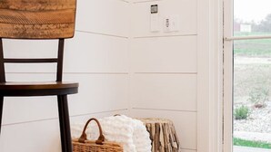 Un rincón de una habitación con una silla de madera, una cesta de mimbre y un pouf blanco frente a una pared blanca con paneles.