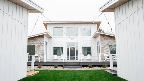 A modern two-story house with a stone facade, a covered patio with white chairs, and a well-maintained lawn.