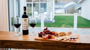 A wooden table with a bottle of wine, two glasses of red wine, and a charcuterie board with fruits, cheese, and crackers.