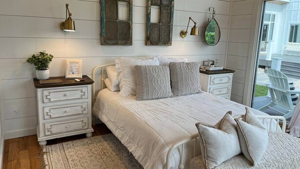 Un dormitorio con una cama grande, paredes blancas, paneles de madera, un aparador, lámparas de noche y vistas al exterior.