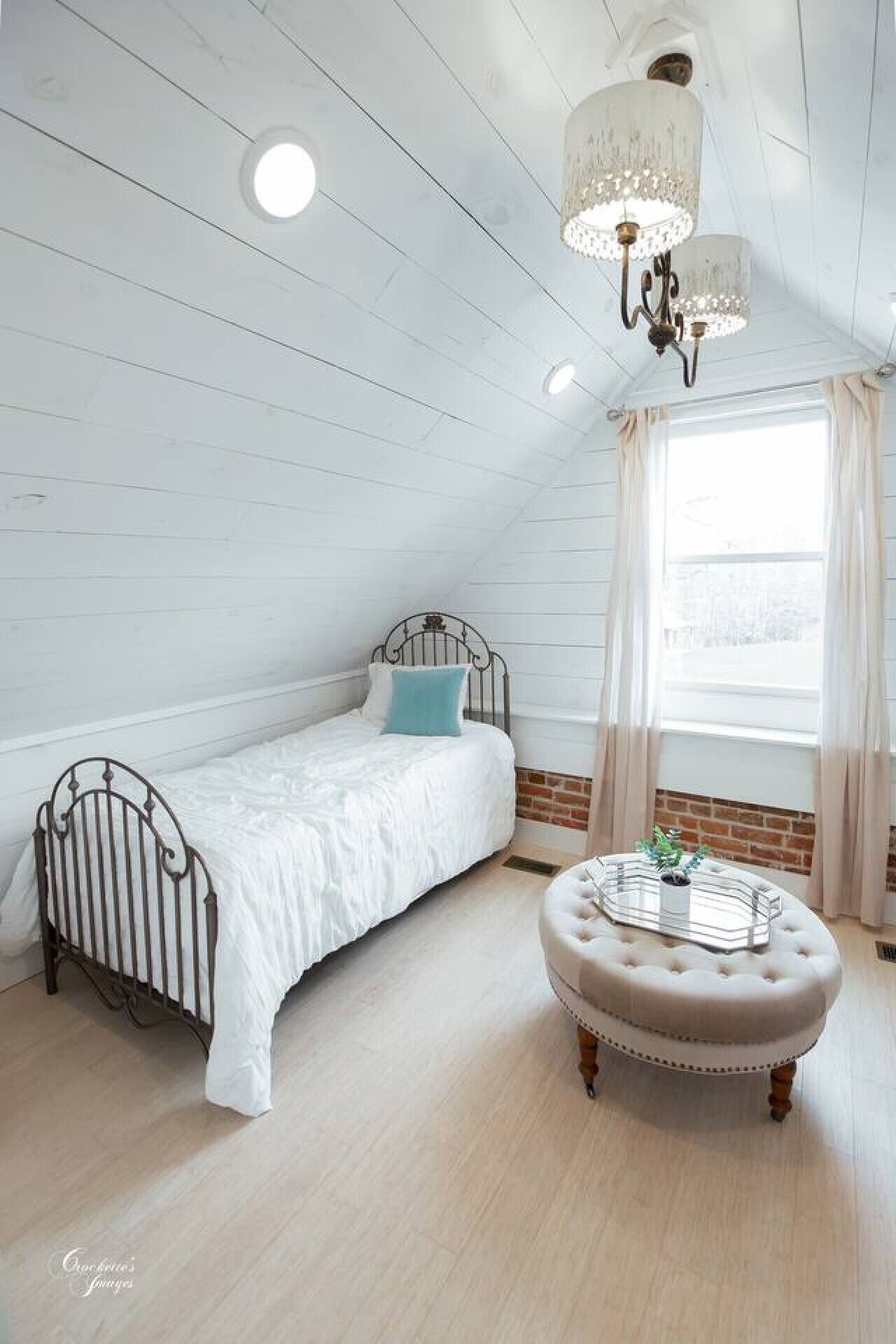 Une chambre avec un lit simple, un pouf rond, un lustre et une fenêtre avec des rideaux.