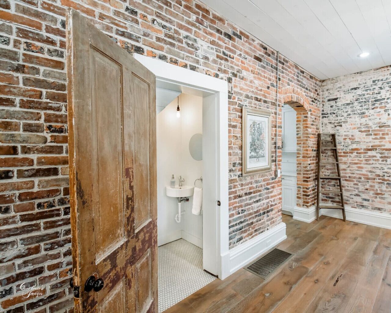 Une pièce aux murs en briques apparentes, avec une porte en bois, un évier blanc et une échelle.