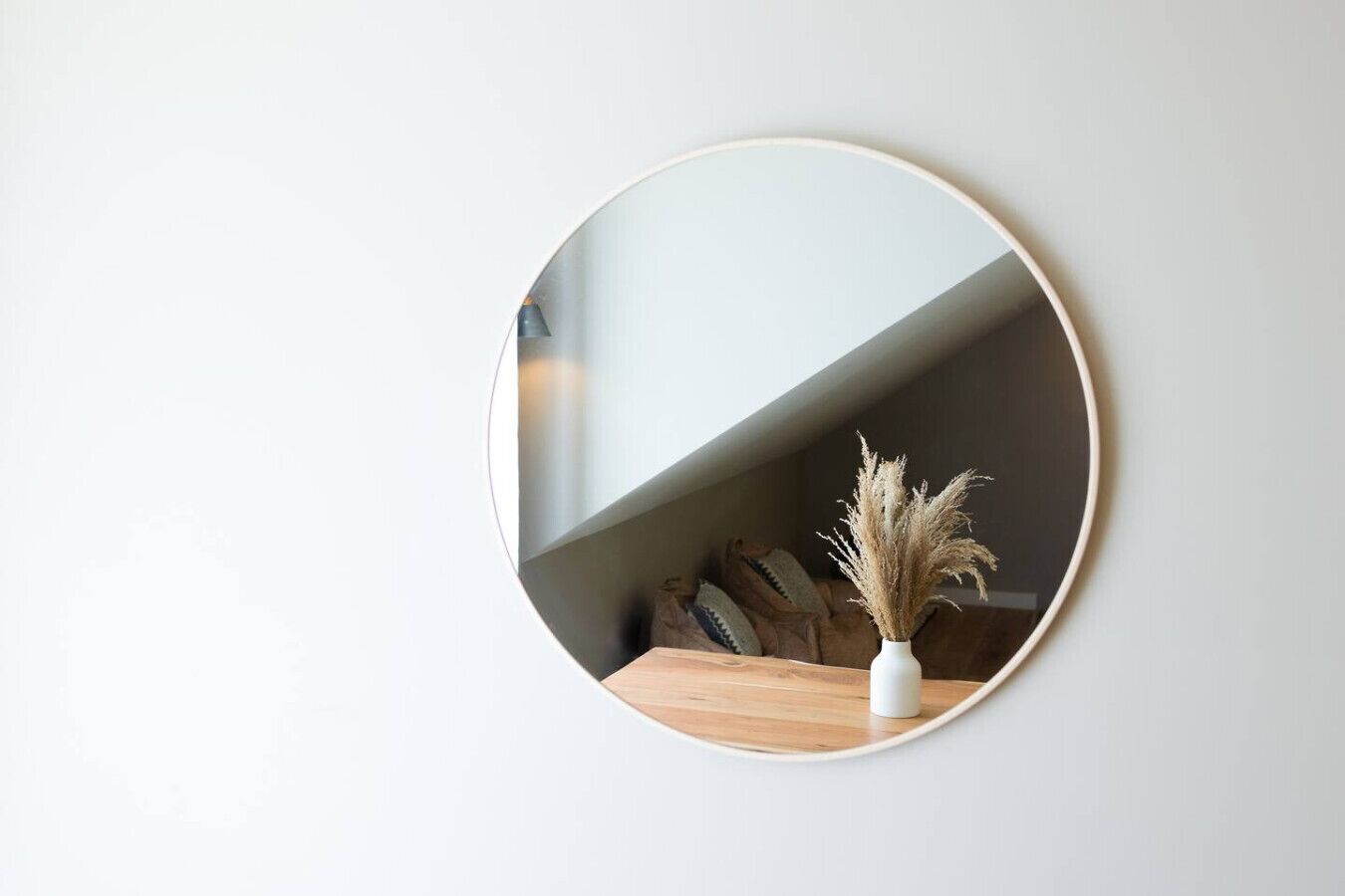 Un miroir rond accroché au mur, avec une table en bois en dessous, un vase contenant des plantes séchées et un canapé avec des coussins à l’arrière-plan.