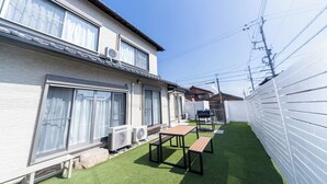 Edificio residencial con patio amoblado, que incluye una mesa, bancos, unidades de aire acondicionado y una cerca blanca.