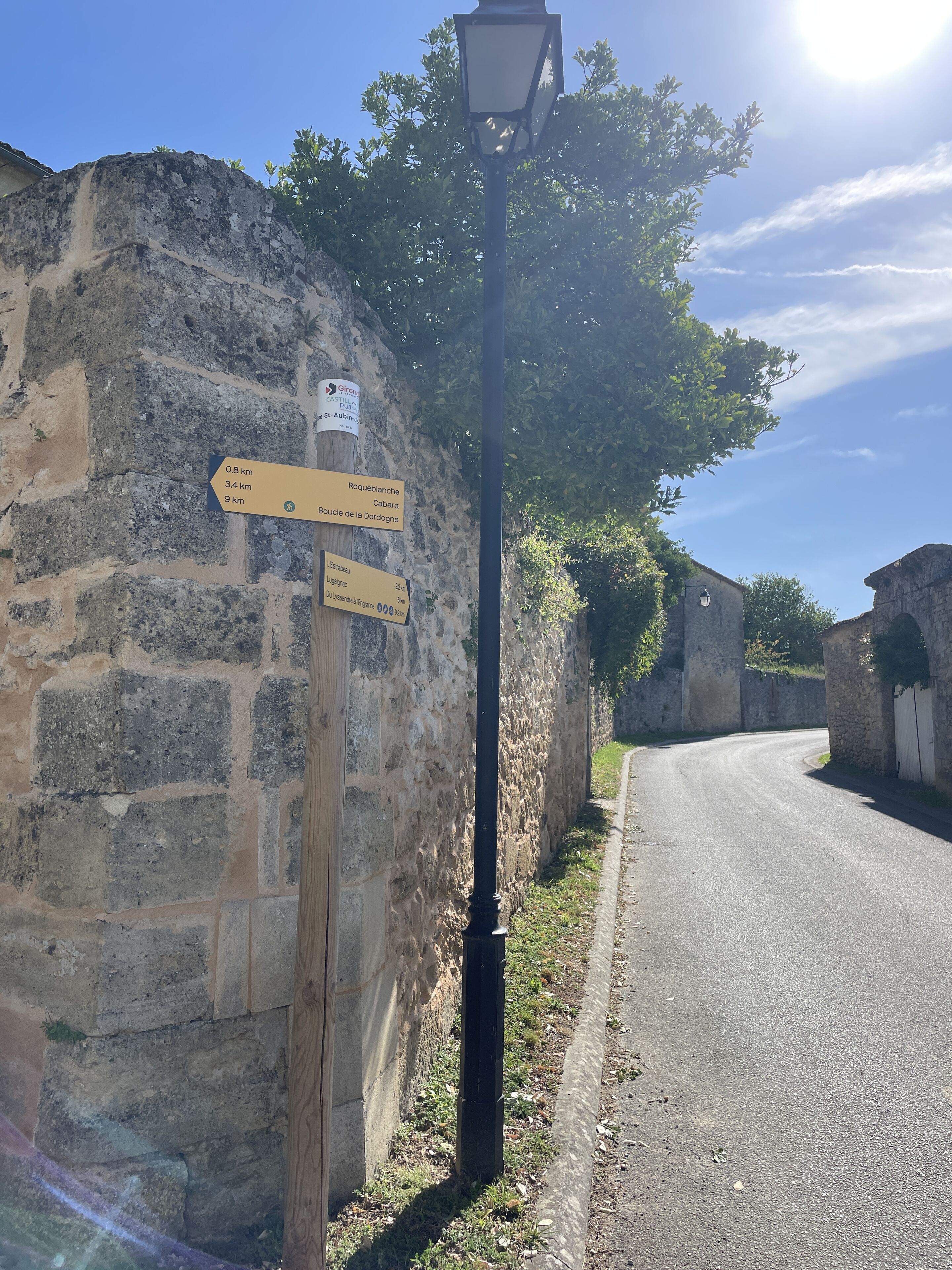 Un mur de pierres avec des panneaux indicateurs, un lampadaire et une route qui tourne vers la droite.