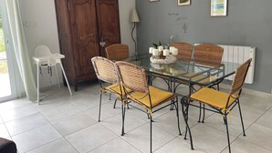 Un área de comedor con una mesa de vidrio rodeada de sillas de mimbre con cojines amarillos, un mueble de madera y cuadros en la pared.