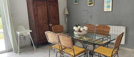 Un área de comedor con una mesa de vidrio rodeada de sillas de mimbre con cojines amarillos, un mueble de madera y cuadros en la pared.