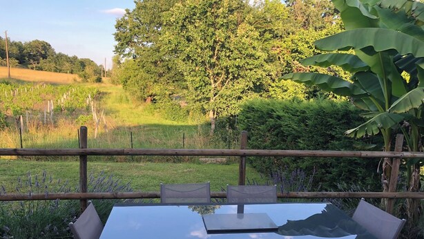 Una terraza de madera con una mesa y sillas de vidrio, rodeada por un cerco de madera, con vistas a un campo cubierto de césped y árboles.