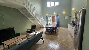 Een ruime woonkamer met een trap, een eettafel met stoelen, een bank en een keuken met een koelkast.