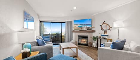 A modern living room with a fireplace, a television, and a view of the outdoors through large windows.