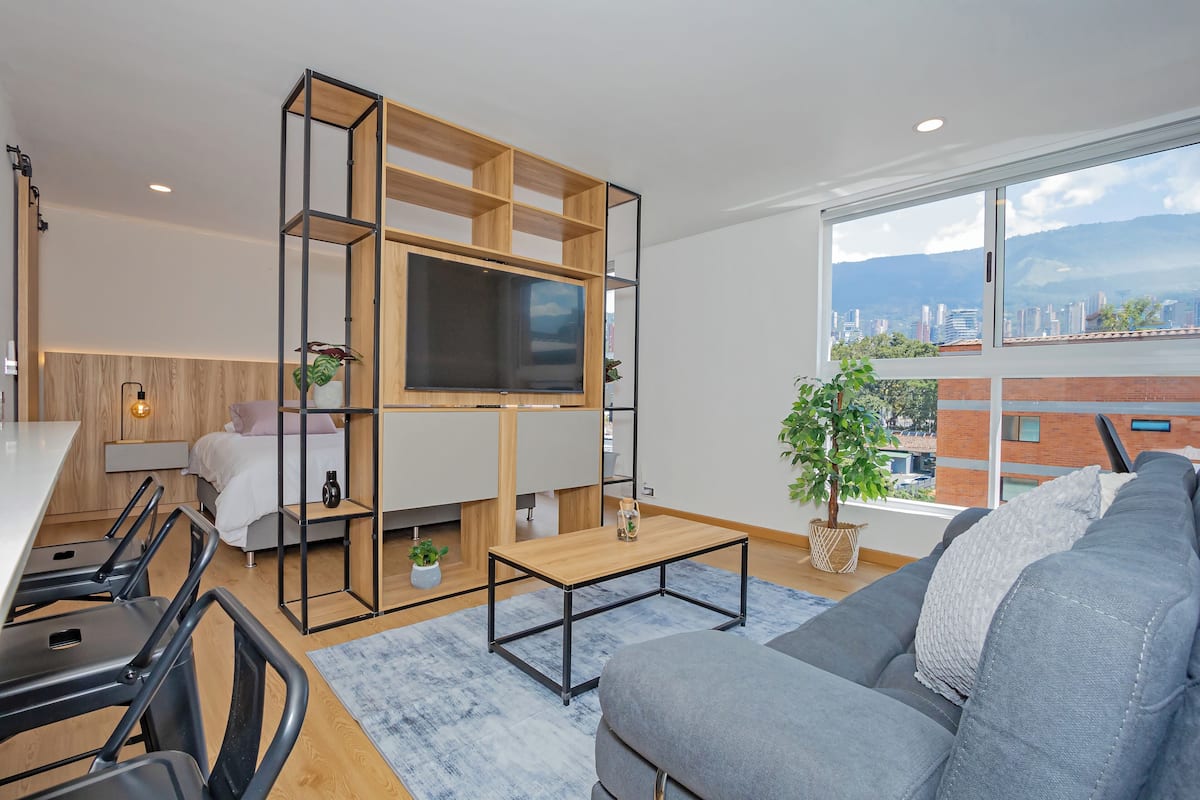 Un salon moderne avec un téléviseur fixé sur un cadre en bois, un canapé gris, un coin repas avec des chaises et une grande fenêtre donnant sur la ville.