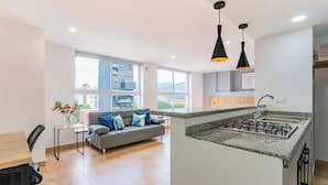 Una cocina moderna con estufa a gas integrada, una zona de comedor con mesa y sillas de madera, un sofá gris con cojines azules, y grandes ventanales con vista a la ciudad.