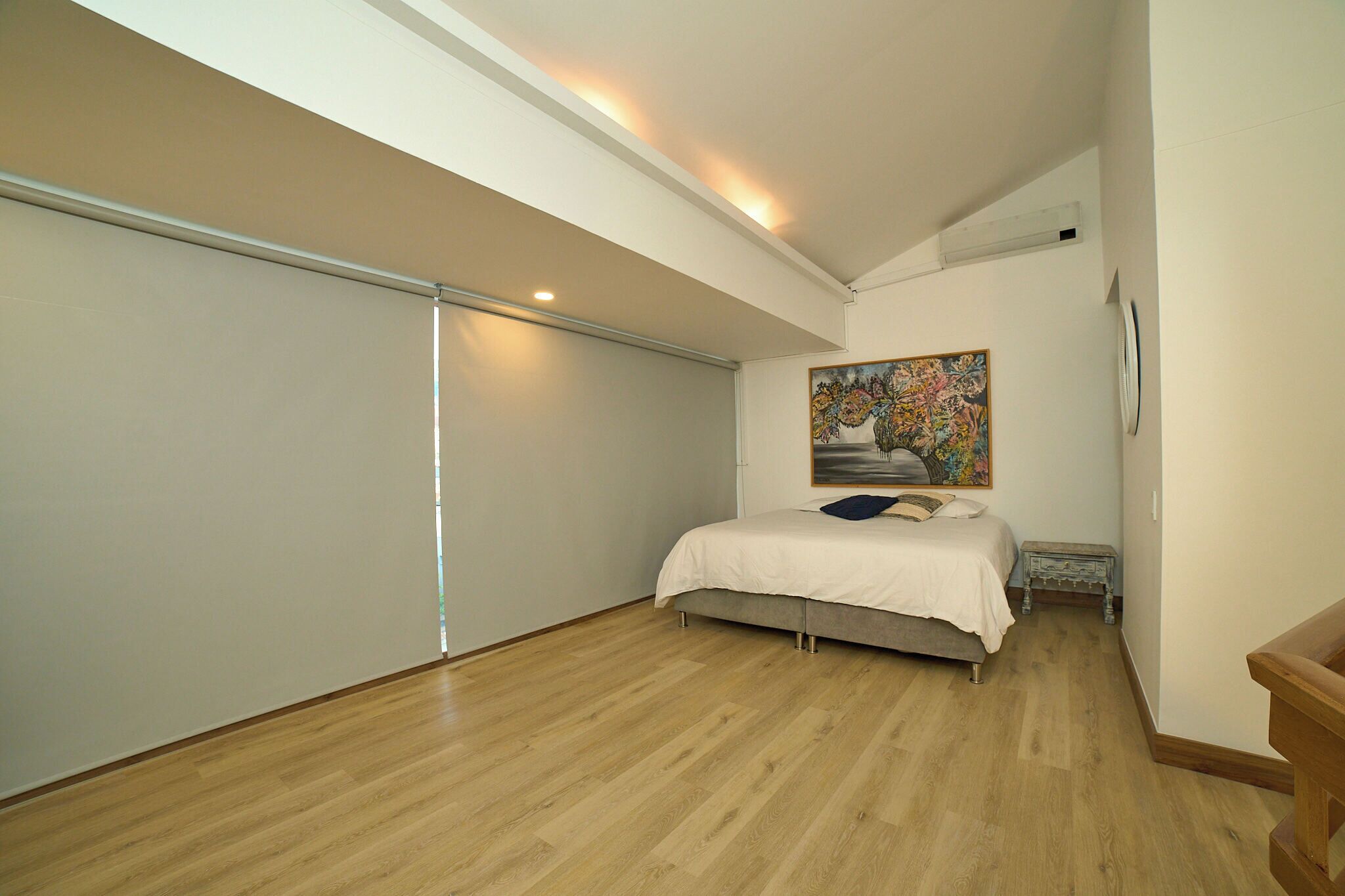 A bedroom with a bed, a painting, a small table, and a wooden floor.
