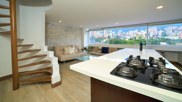 Una cocina moderna con una isla central, una escalera, un sofá y vistas a la ciudad a través de amplios ventanales.