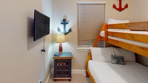 Une pièce avec un téléviseur fixé au mur, un lit superposé en bois, une fenêtre avec des stores et une décoration en forme d’ancre sur le mur.
