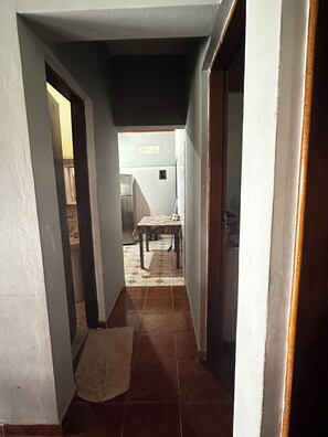 A narrow hallway with a tiled floor, a table with a patterned tablecloth, and a refrigerator in the background.