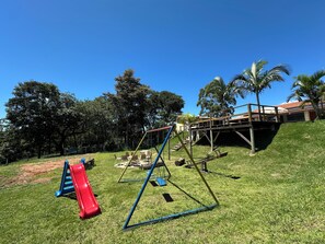A playground with a slide and swings on a grassy hill with trees and a building in the background. - Naldinho's Farm (Mairinque)