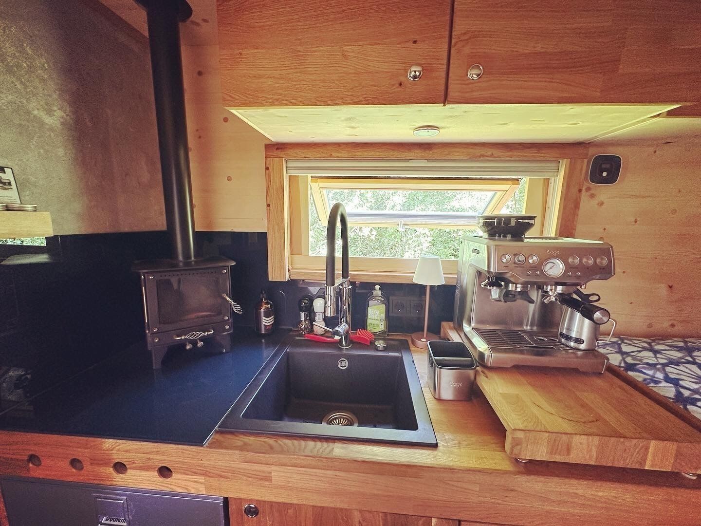 A compact kitchen with a wood stove, a coffee machine, and a window providing natural light.