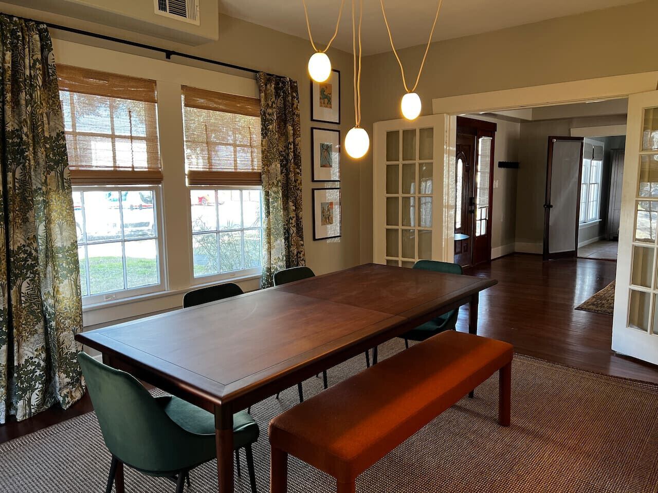 A spacious dining area with a long wooden table, green chairs, and a bench. There are large windows with curtains, a refrigerator, and a door leading to another room.