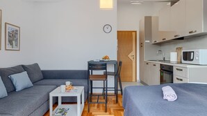 Un espacio habitable compacto con sofá, una pequeña mesa de comedor, una zona de cocina con microondas y una cama con una toalla enrollada.