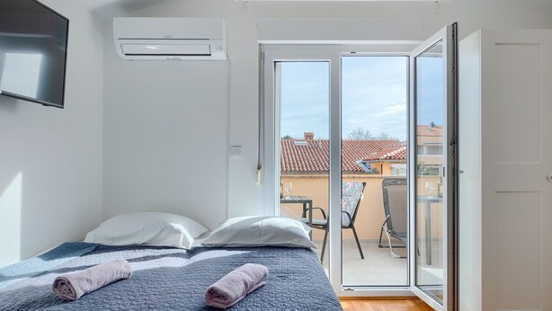 Un dormitorio con cama, almohadas, una toalla enrollada y televisor. Hay una puerta corrediza de vidrio que da a un espacio exterior con mesa y sillas.