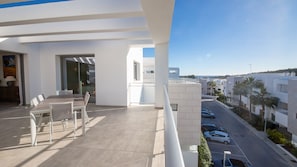 Un balcón con una mesa y sillas para comer, vista a una calle con autos y edificios, y cielo despejado.