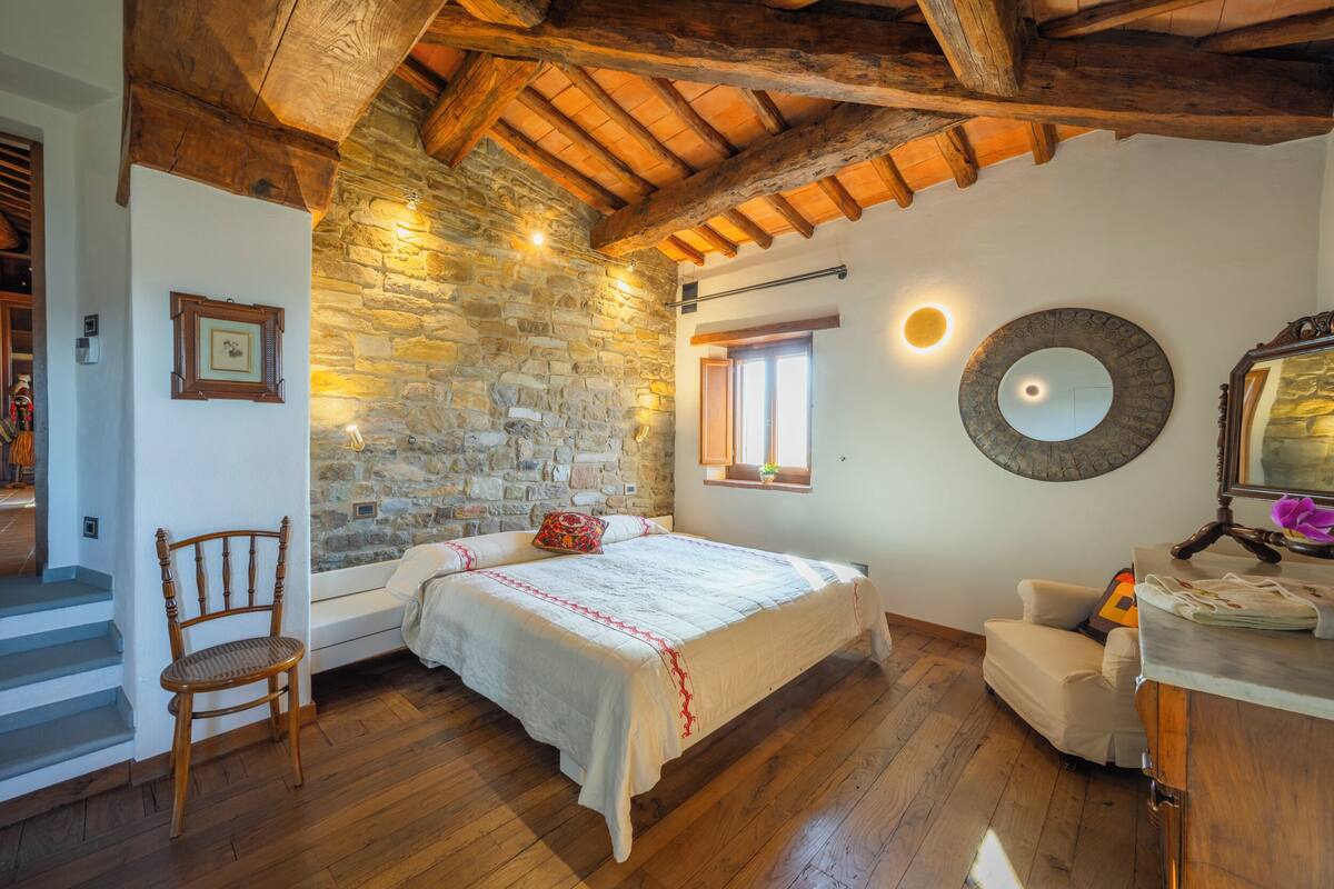 A bedroom with a stone wall, wooden beams, a bed, a chair, and a desk.
