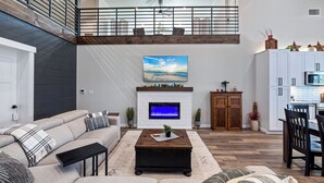 Un salón amplio con techo alto, chimenea, televisor grande y comedor con armarios y sillas de madera.