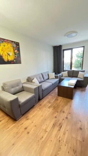 Ein modernes Wohnzimmer mit einem grauen Ecksofa, einem Holztisch und einem großen Fenster, das für natürliches Licht sorgt.