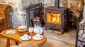 A cozy room with a stone wall, a wooden table with a dessert, tea set, and a fireplace with logs.