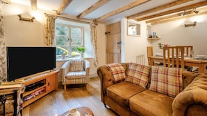 A cozy living room with a television, a plaid armchair, a sofa with plaid cushions, a wooden dining table, and a window with curtains.