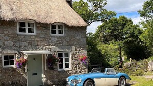 A stone cottage with a thatched roof, a vintage blue and white car parked in front, and a sign indicating 'The Cottage' above the entrance.