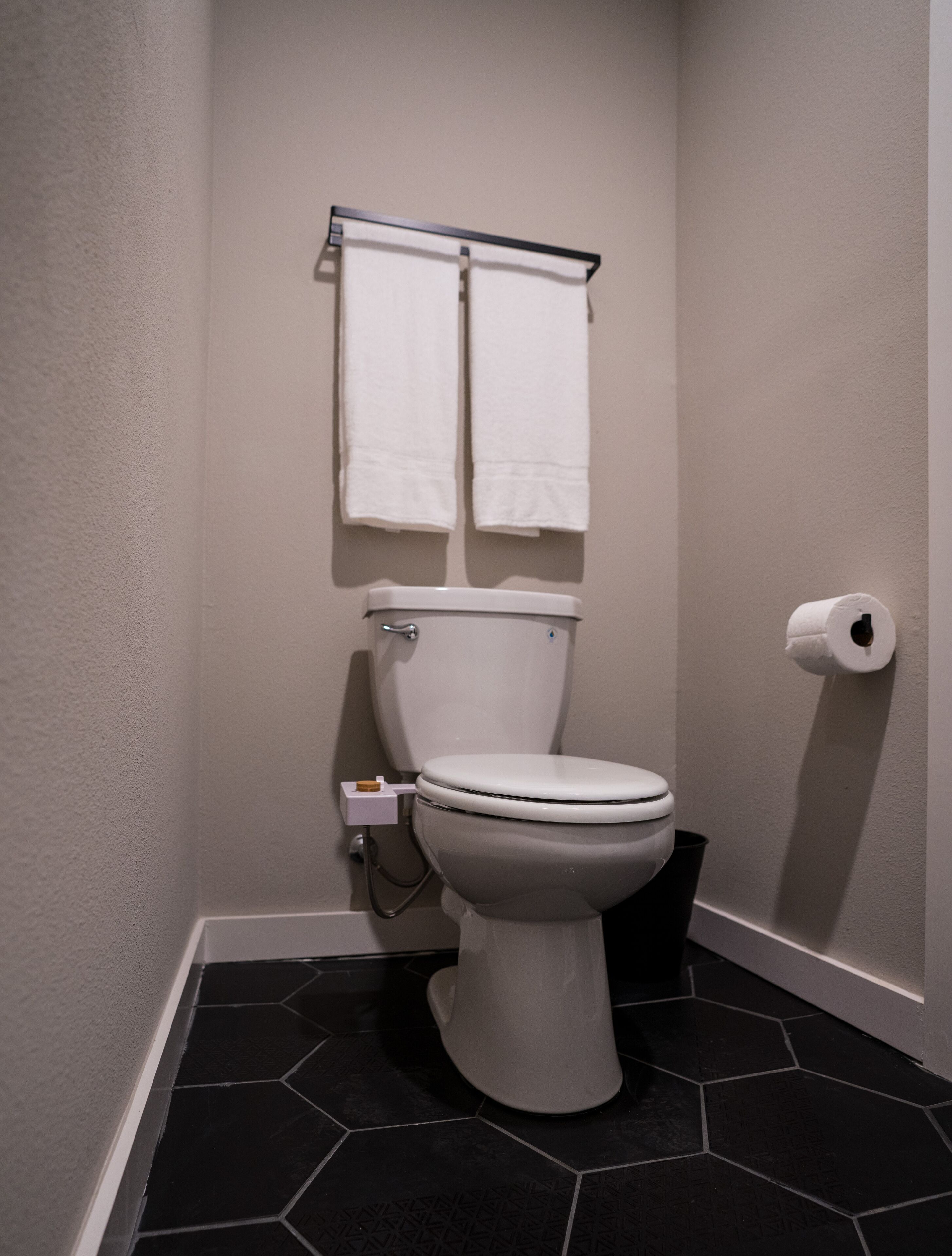 Un baño pequeño y moderno con inodoro, un dispensador de papel higiénico y un perchero con dos toallas blancas.