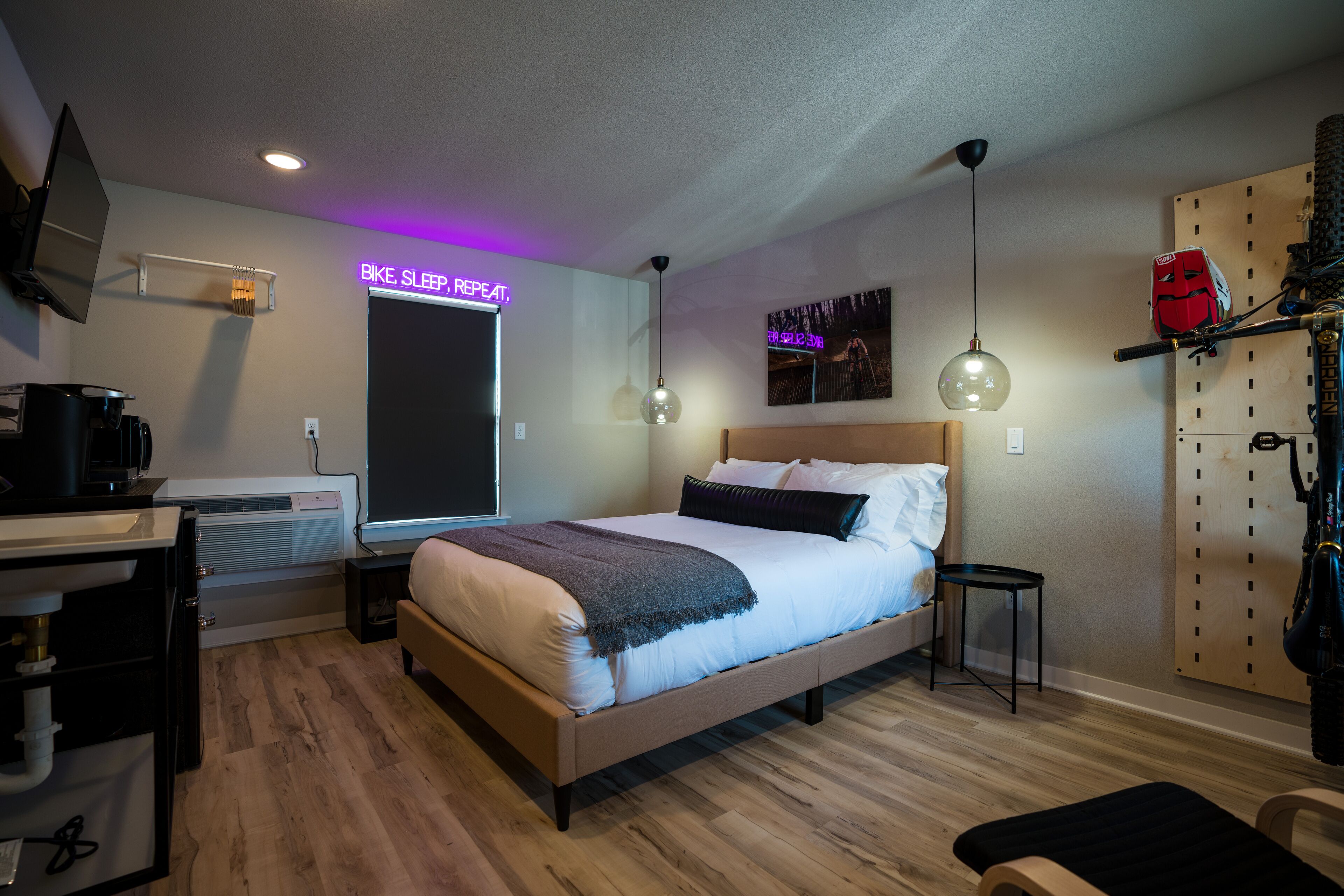 A modern bedroom with a bed, bedside tables, a desk with a computer, a wall-mounted TV, and a neon sign.
