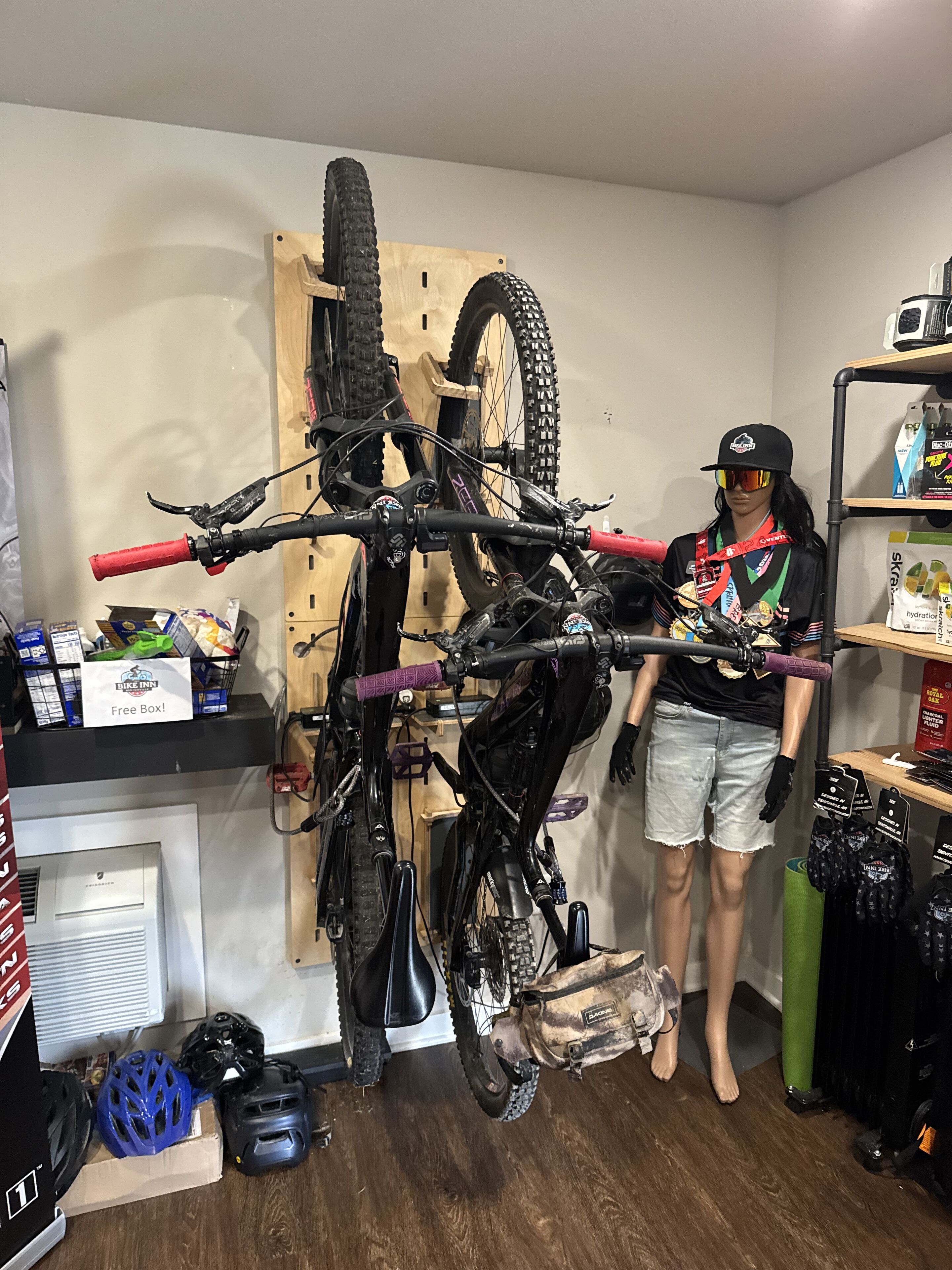 Une pièce dans laquelle on peut voir un vélo fixé sur un support en bois, un mannequin vêtu d’une tenue de cycliste, et divers accessoires de cyclisme disposés sur des étagères.