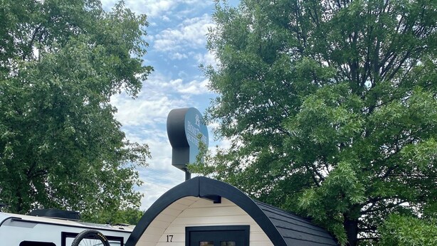 A small, single-story building with a black roof and white walls, featuring a door with a neon sign that reads "Bike Sleep Repeats." There are two black chairs outside and a bicycle parked to the left. - The Bike Inn (Bentonville)