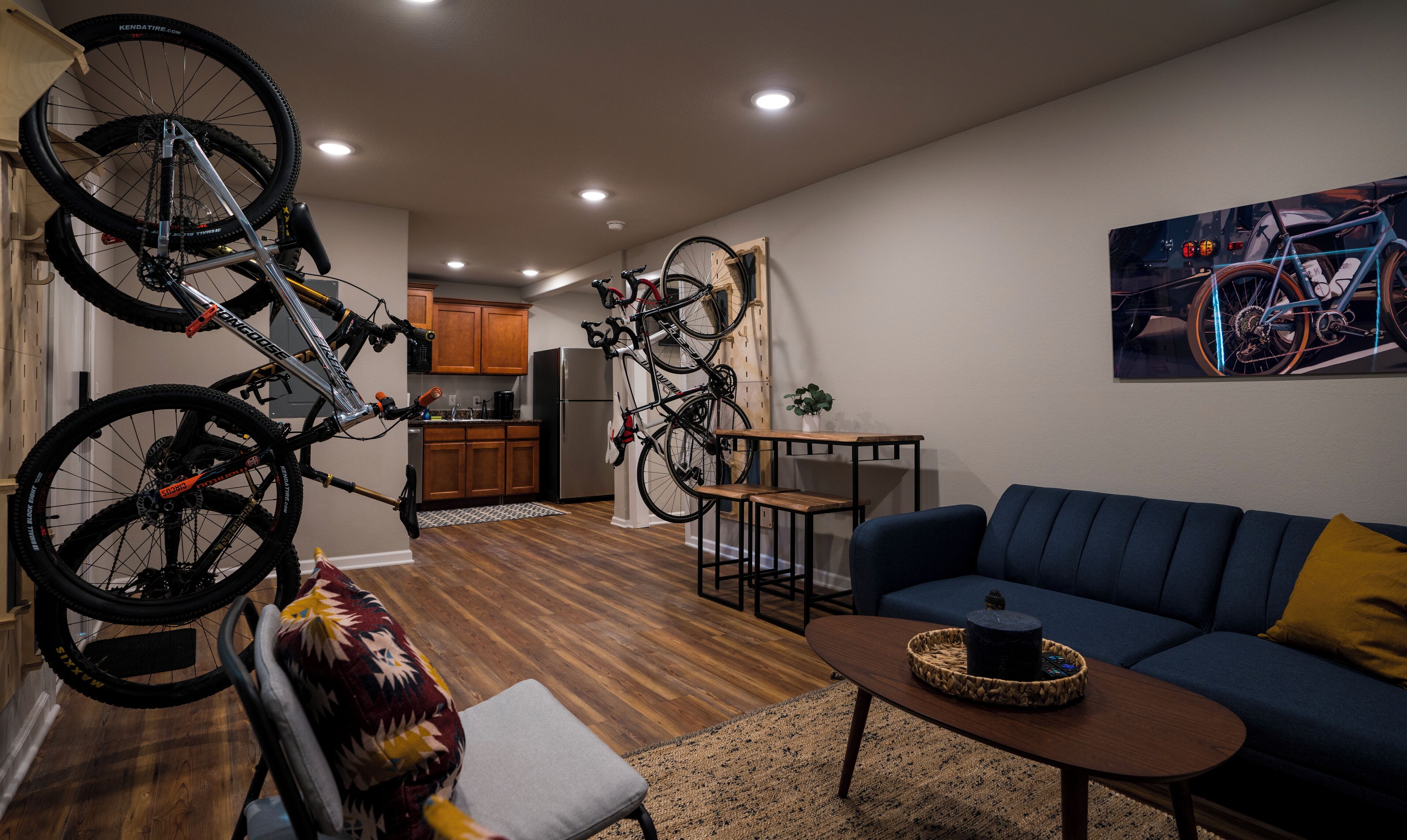 A living room with a blue sofa, a coffee table, and a dining table. There are bicycles mounted on a wall and a kitchen in the background.