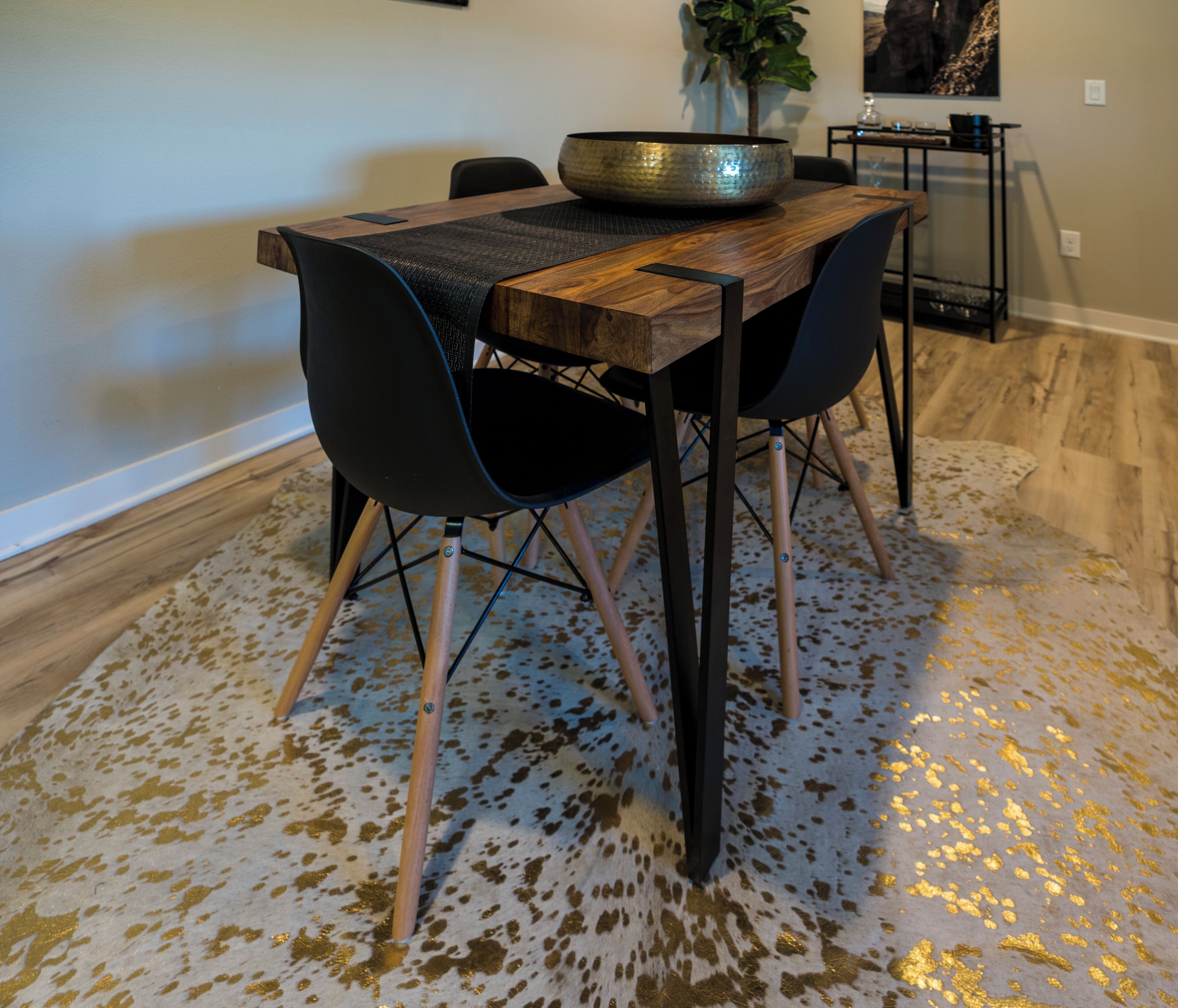 Un coin repas avec une table en bois, deux chaises noires, un grand saladier et un tapis à motifs.