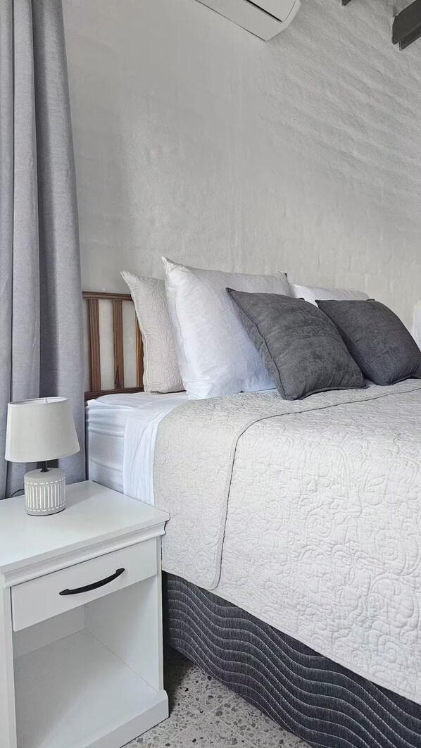 Un dormitorio ordenado con cama, almohadas, una mesita de noche con lámpara y un mueble auxiliar.