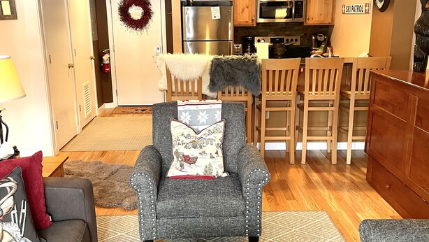 A modern living room with a grey armchair, a wooden coffee table with a vase of flowers, a grey sofa, and a dining area with wooden chairs and a kitchen in the background. - Ski in/out @Base Lodge at  Clock tower,Okemo. Building B  Ground Level sleeps 6 (Ludlow)