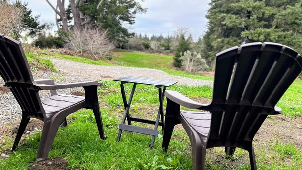 Dos sillas Adirondack y una mesa pequeña sobre un sendero de grava, rodeado de césped, árboles y follaje.