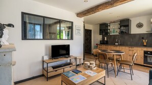 Ein modernes Wohnzimmer mit Fernseher, Couchtisch und Sesseln. Im Hintergrund ist eine Küche mit Essbereich erkennbar.