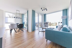 A modern living room with a sofa, chairs, a dining table, and large windows.