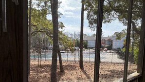 A view through a window of a fenced outdoor area with trees, a building, and a parked truck. - Anchored At Last 3-bed Condo Kill Devil Hills w/ Large Deck and View of the Pool (Kill Devil Hills)