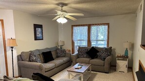 A living room with two sofas, a coffee table, a ceiling fan, and a window with lace curtains. - Anchored At Last 3-bed Condo Kill Devil Hills w/ Large Deck and View of the Pool (Kill Devil Hills)