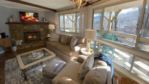 A cozy living room with a stone fireplace, a large window with a view of trees, a sofa with patterned cushions, and a chandelier. - Sugar's Edge Luxury Mountain Treehouse with AirCon (Sugar Mountain)