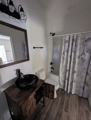 Une salle de bain avec un meuble-lavabo en bois, un lavabo noir, des toilettes blanches, une douche avec un rideau et un miroir au mur.